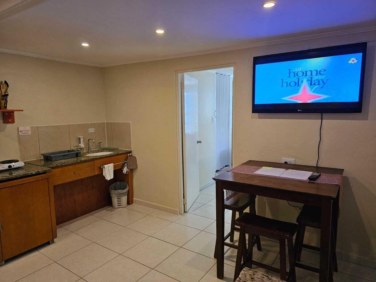 Une cuisine compacte avec un coin repas, un téléviseur diffusant le message « votre maison, vos vacances », et une porte donnant sur une autre pièce.