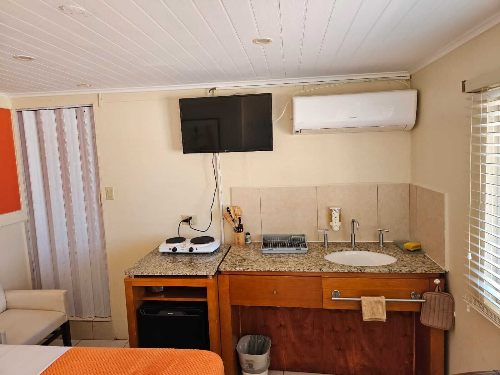 A compact hotel room with a kitchenette, a sink, a mirror, a television mounted on the wall, and a window with blinds.
