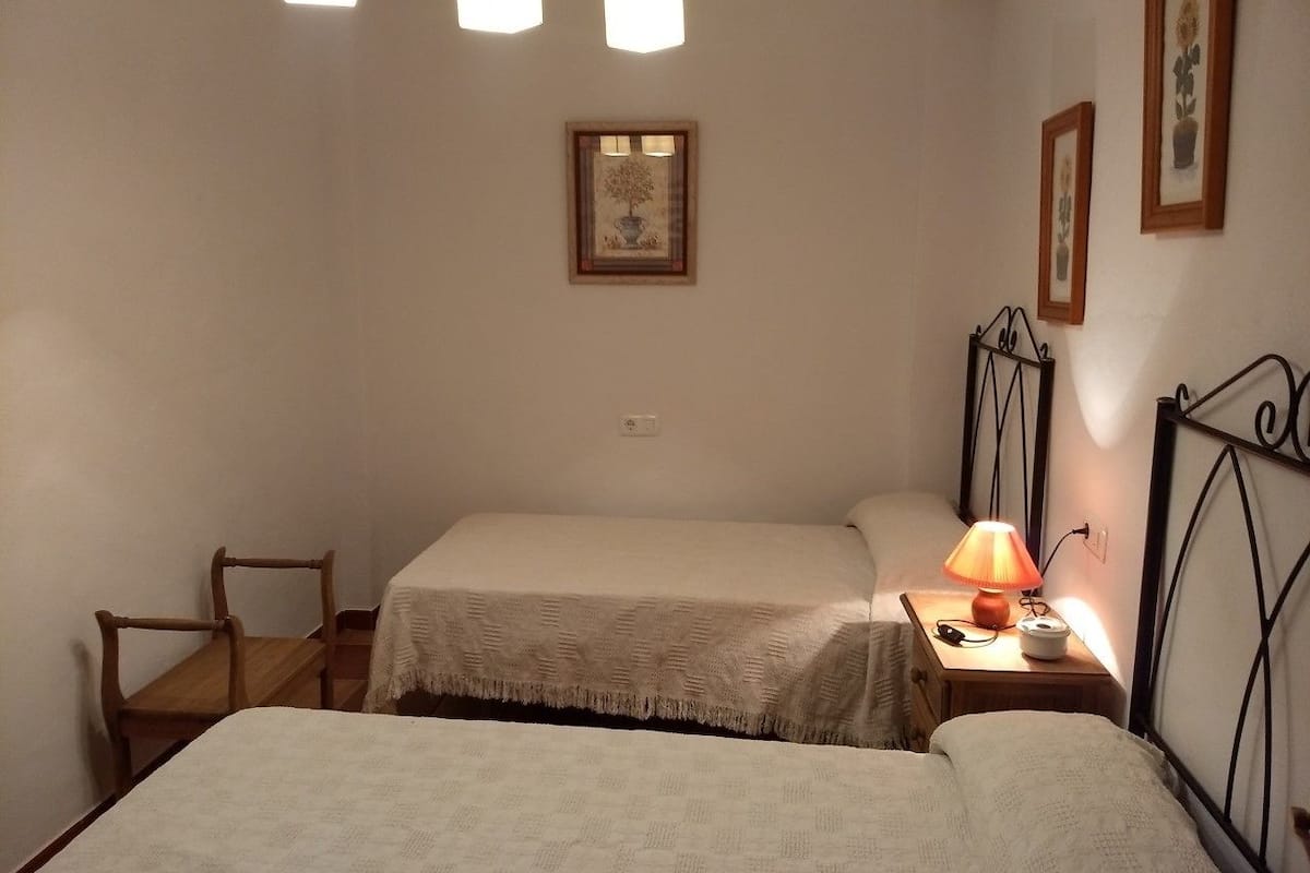 A small, cozy bedroom with two single beds, a wooden chair, a nightstand with a lamp, and framed pictures on the wall.