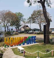 Letrero con la palabra 'HONDURAT' en letras grandes y coloridas, rodeado de árboles y barcos, que indica una ubicación frente al agua.