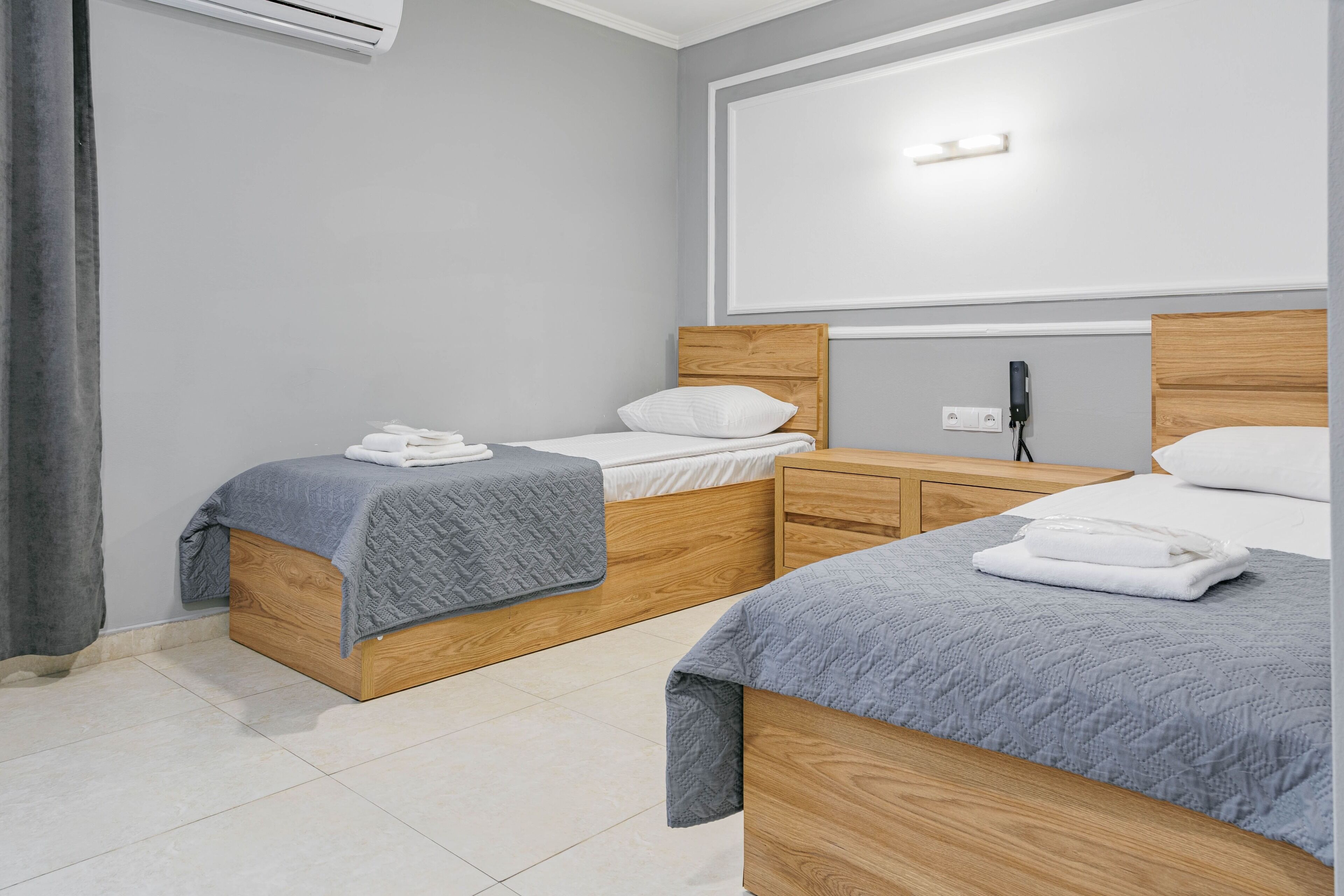 Camera d'albergo con due letti in legno, biancheria grigia, un comodino e un condizionatore d'aria a parete, oltre a una grande finestra.