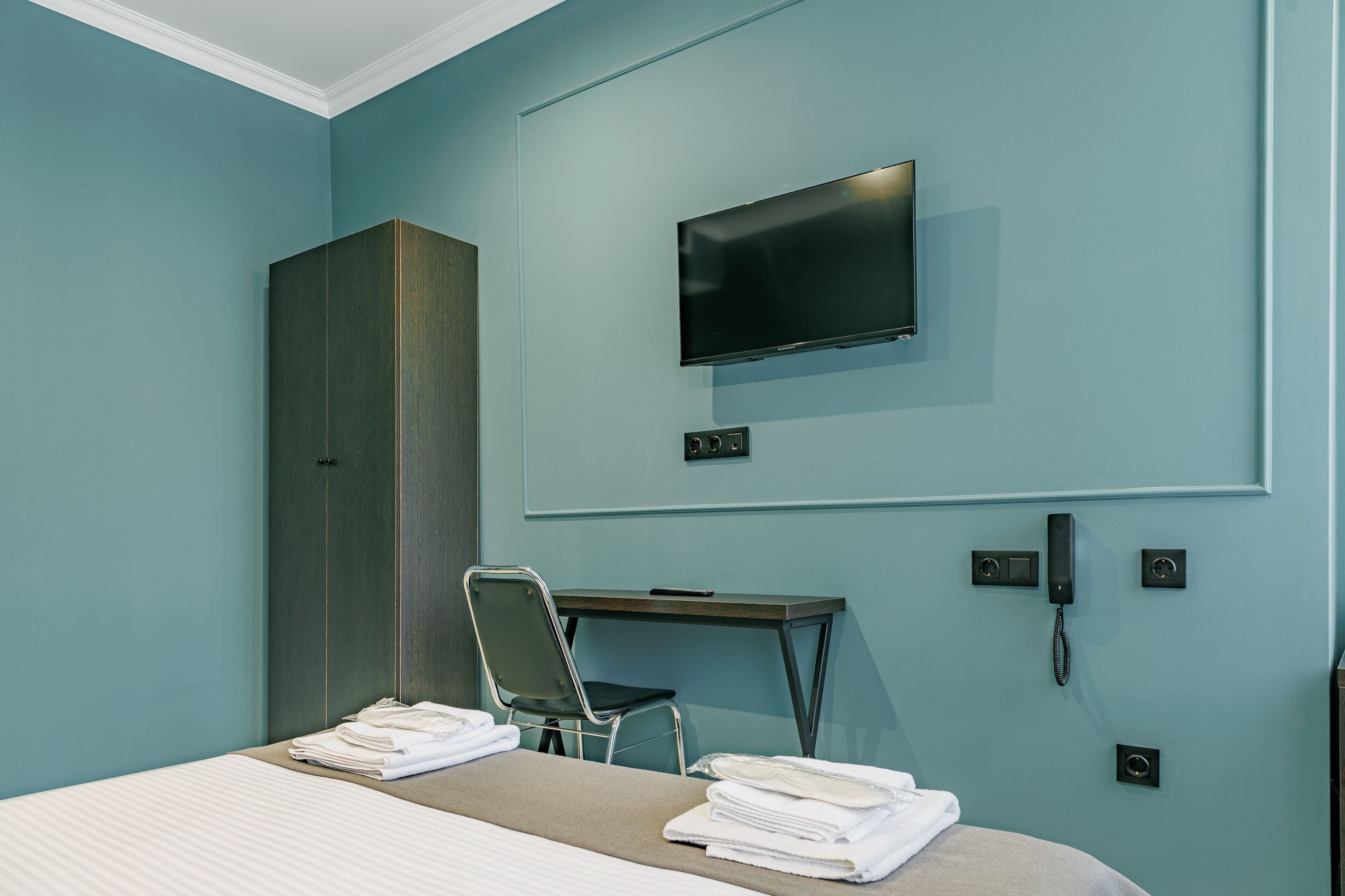 A hotel room with a bed, a desk, a chair, a wardrobe, a flat-screen TV, and a telephone.