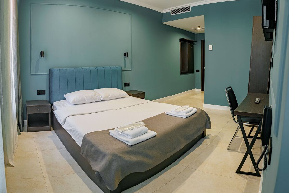 A modern hotel room with a large bed, bedside tables, a desk with a chair, and a mirror.