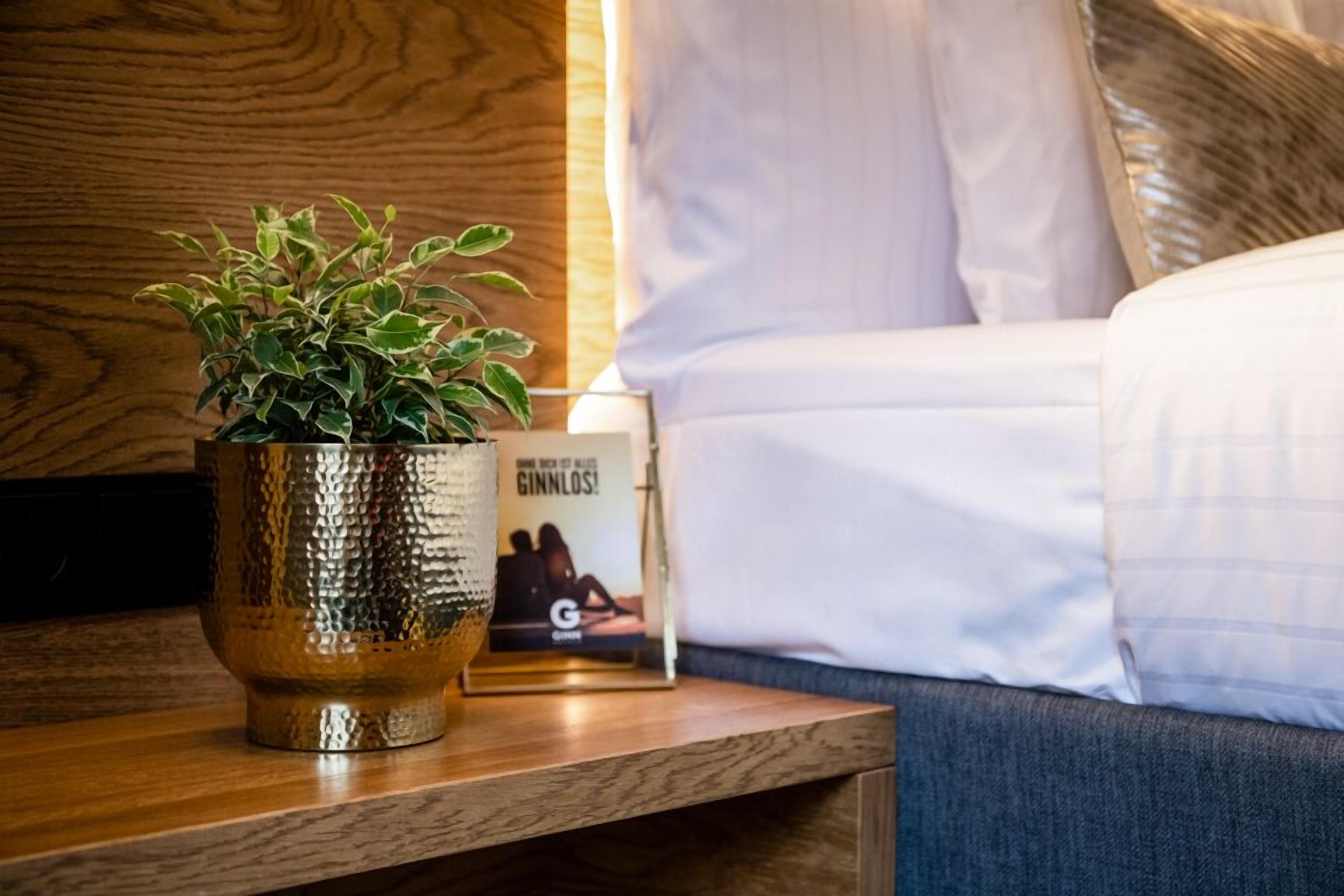 A wooden nightstand with a textured metallic vase containing a green plant, a framed picture, and a lamp.