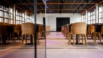 A conference room with rows of wooden chairs and tables, large windows on the sides, and a modern ceiling design.