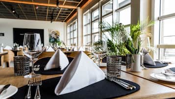 Een restaurantscène met gedekte tafels, zwarte placemats, wijnglazen en een potplant.