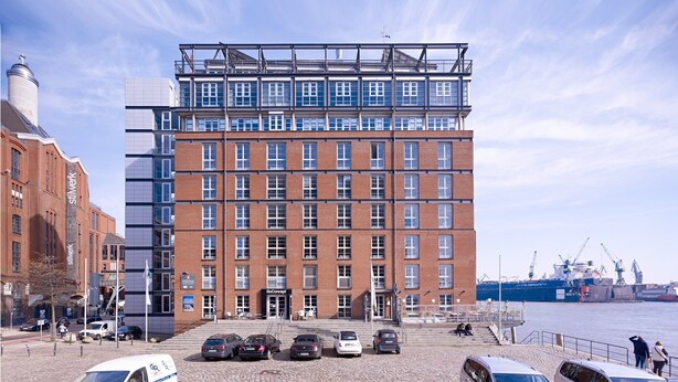 Un edificio moderno con fachada de ladrillo y varias ventanas, situado cerca de una orilla con un barco grande en el fondo.
