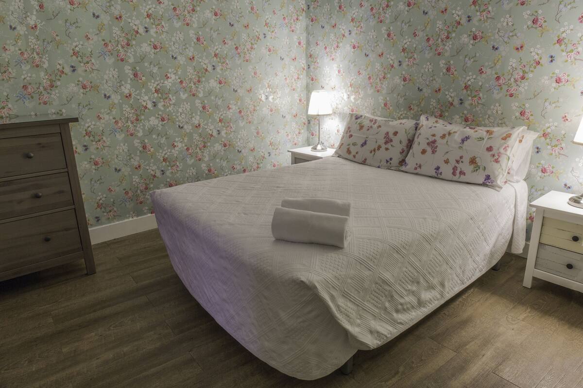 A neatly made bed with a white bedspread and pillows, a wooden dresser, and a nightstand with a lamp.