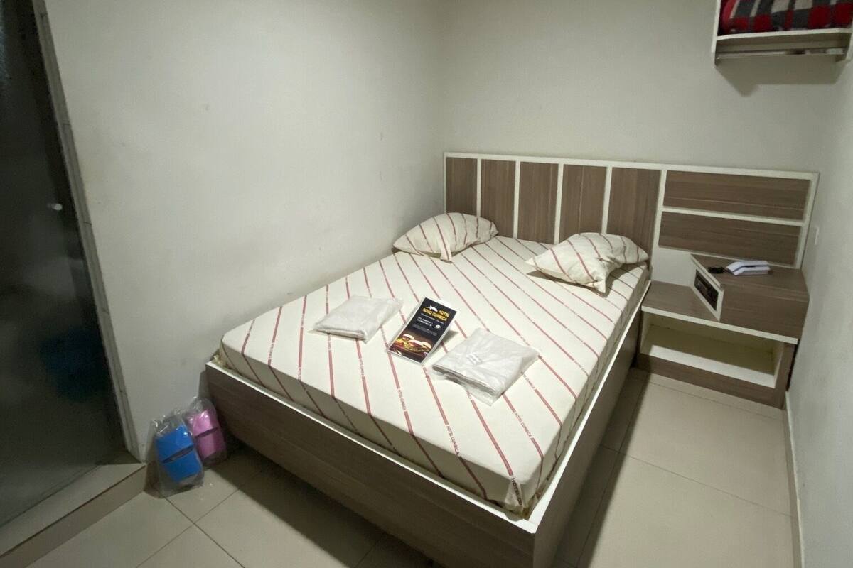Un lit bien fait, recouvert d’une couvre-lit à rayures, une table de chevet avec une lampe et une petite étagère sur laquelle se trouve un objet à carreaux rouges et blancs.