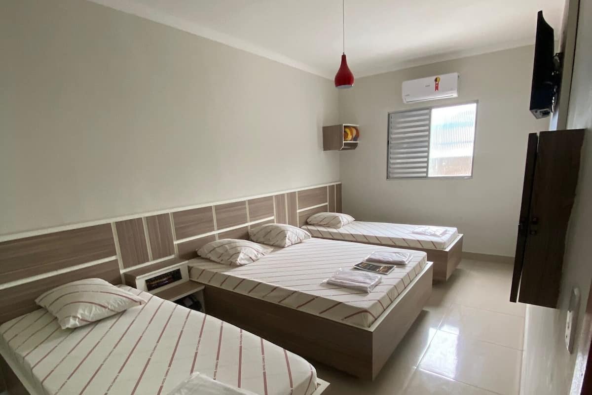 Une chambre d’hôtel avec deux lits, un lampadaire suspendu rouge, une petite étagère avec un tableau, et une fenêtre avec des stores.
