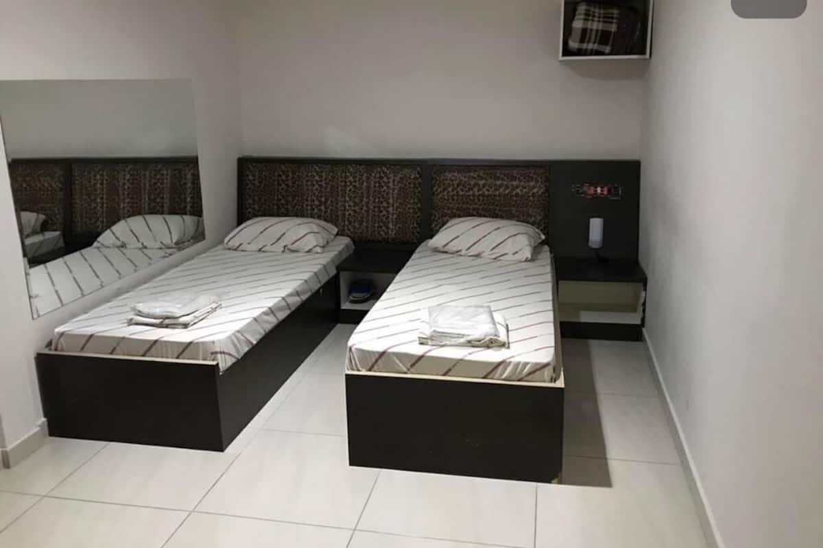 Une chambre avec deux lits, une petite table, une chaise, une table de chevet et une étagère fixée au mur.