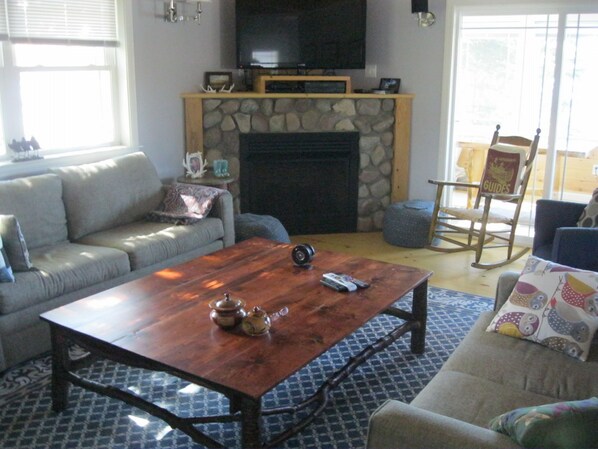 A living room with a stone fireplace, a sofa, a coffee table, and a television mounted above the fireplace. - New Beam construction house on pristine Back Lake (Pittsburg)