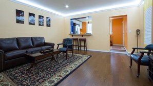 A living room with a dark leather sofa, a coffee table, and a patterned rug. There are two chairs and a small table with a mirror above it. The room has wooden flooring and beige walls.