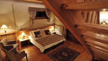 A cozy bedroom with a wooden staircase, a bed with a plaid bedspread, two bedside lamps, a window with curtains, and a patterned rug.
