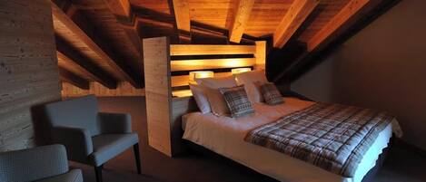 A cozy attic bedroom with a wooden ceiling, a bed with a plaid bedspread, and two upholstered chairs.