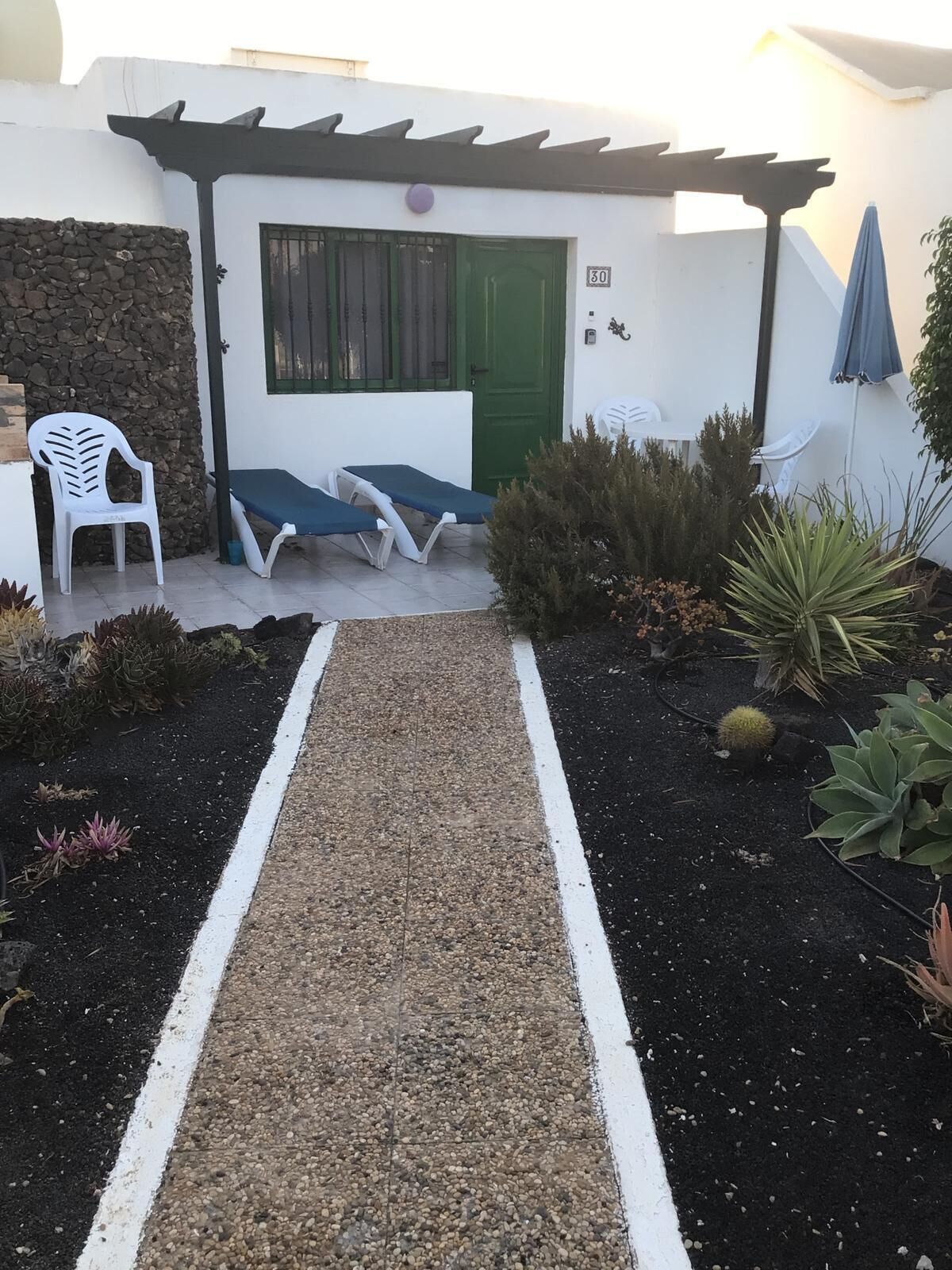Een pad dat leidt naar een gebouw met een groene deur, twee lounge stoelen en een witte stoel buiten.