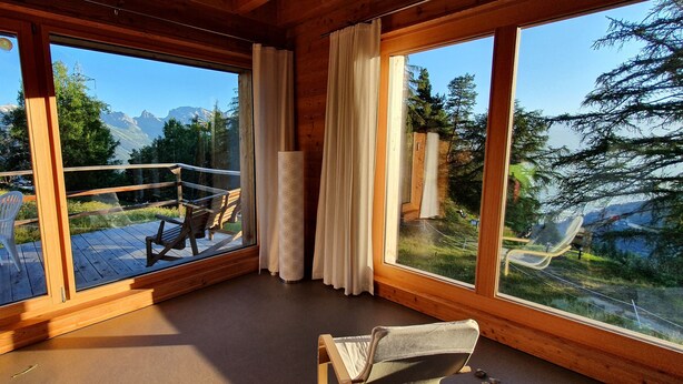 Ein Zimmer mit großen Fenstern und Blick auf die Berge, eine Holzterrasse davor und ein Stuhl im Inneren.