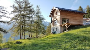 Eine Holzhütte mit Balkon, umgeben von üppigem Grün und hohen Bäumen, mit Blick auf eine Berglandschaft.