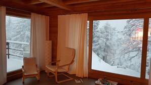 Ein gemütliches Zimmer mit Holzbalkendecke, einem großen Fenster mit Vorhängen, einem Sessel und einem kleinen Tisch mit Tablett.