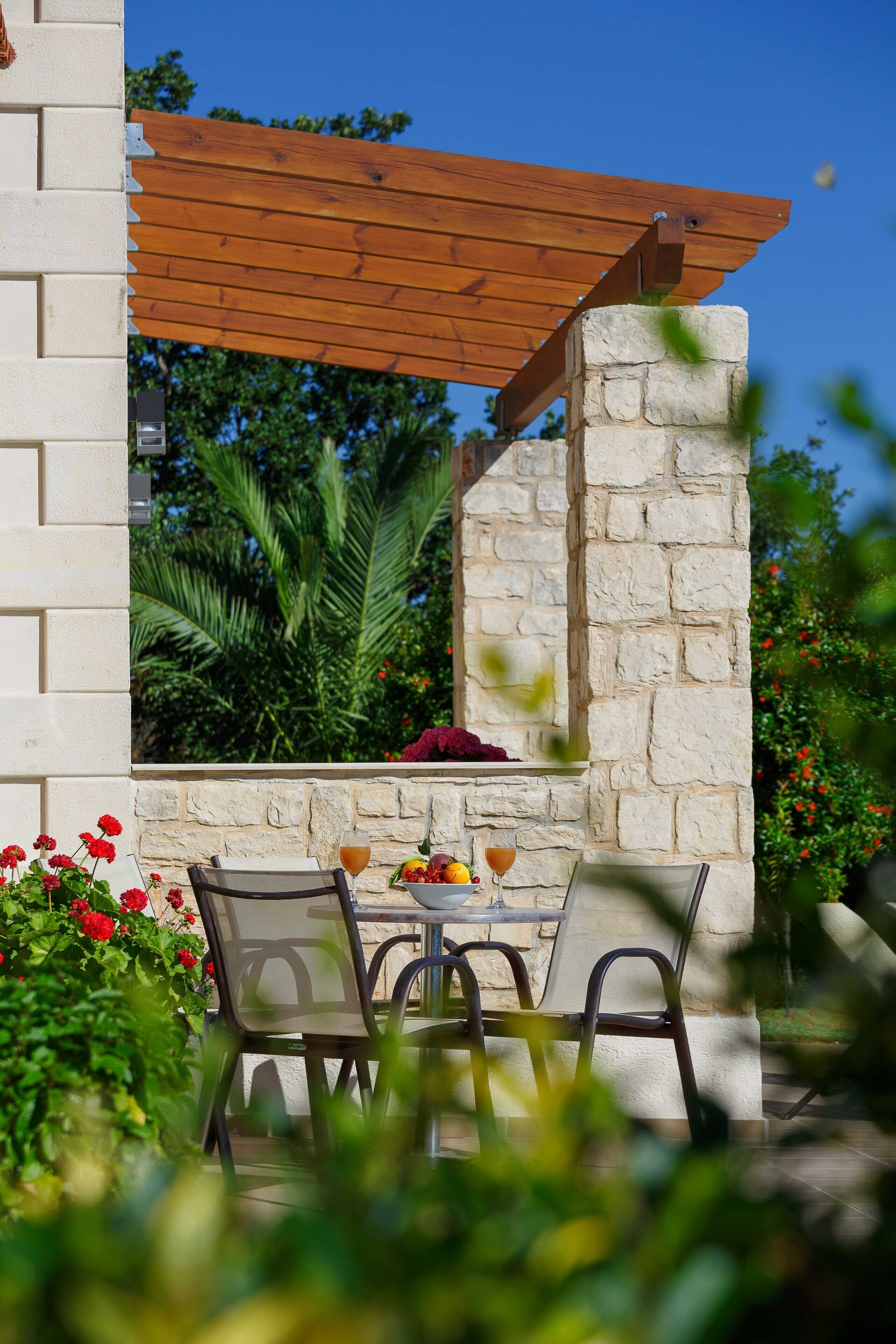 An outdoor patio with a stone wall, wooden pergola, and a table with chairs.