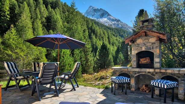 Un área de estar al aire libre con mesas y sillas de piedra, una chimenea de piedra y vistas a una cadena montañosa boscosa.