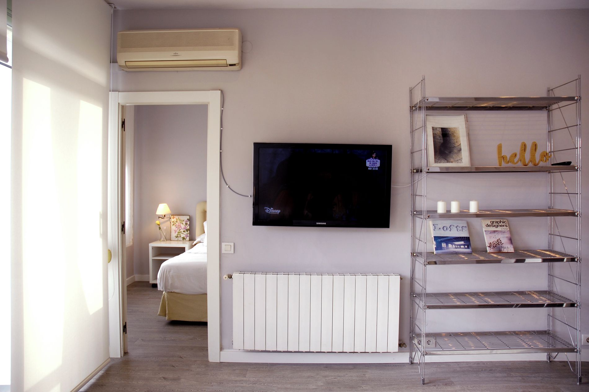 A hotel room with a bed, a TV mounted on the wall, a radiator below it, and a shelving unit with decorative items.