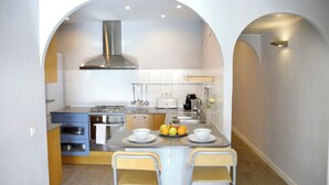 Une cuisine compacte équipée d’une cuisinière à gaz, d’un four et d’appareils électroménagers modernes. On y trouve deux chaises autour d’une petite table, sur laquelle est posé un panier de fruits.