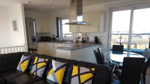 A modern living room with a black leather sofa, a glass dining table, and a kitchen in the background. - 5* Self Catering Penthouse, Llanelli, Beach, Sea Views, Walks, Golf, Cycle. (Llanelli)