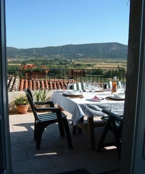 An outdoor dining area with a table set for four, surrounded by terracotta pots and potted plants, overlooking a scenic landscape with hills.