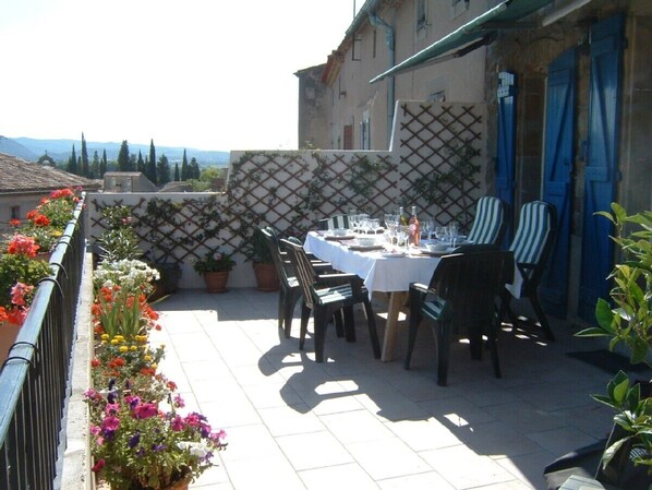 An outdoor dining area with a table set for four, surrounded by chairs. There are potted plants along the railing and a view of the countryside in the background.
