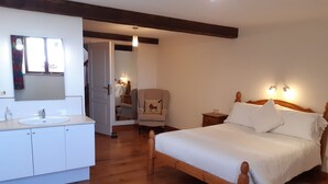 Un dormitorio con cama de madera, ropa de cama blanca, una mesita de noche con lámpara, una silla, suelo de madera, un armario blanco y un baño con lavabo y espejo.