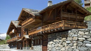 Eine Holzhütte mit Balkonen, Steinmauern und einem Bergpanorama im Hintergrund.