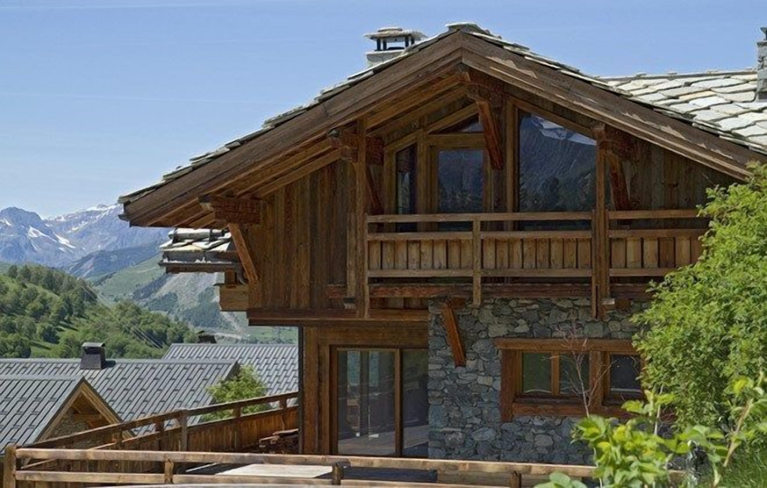 Eine Holzhütte mit Steinunterbau, großen Fenstern und einem Balkon mit Blick auf eine Berglandschaft.
