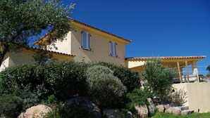 Un bâtiment jaune avec deux fenêtres, entouré de verdure et de rochers.