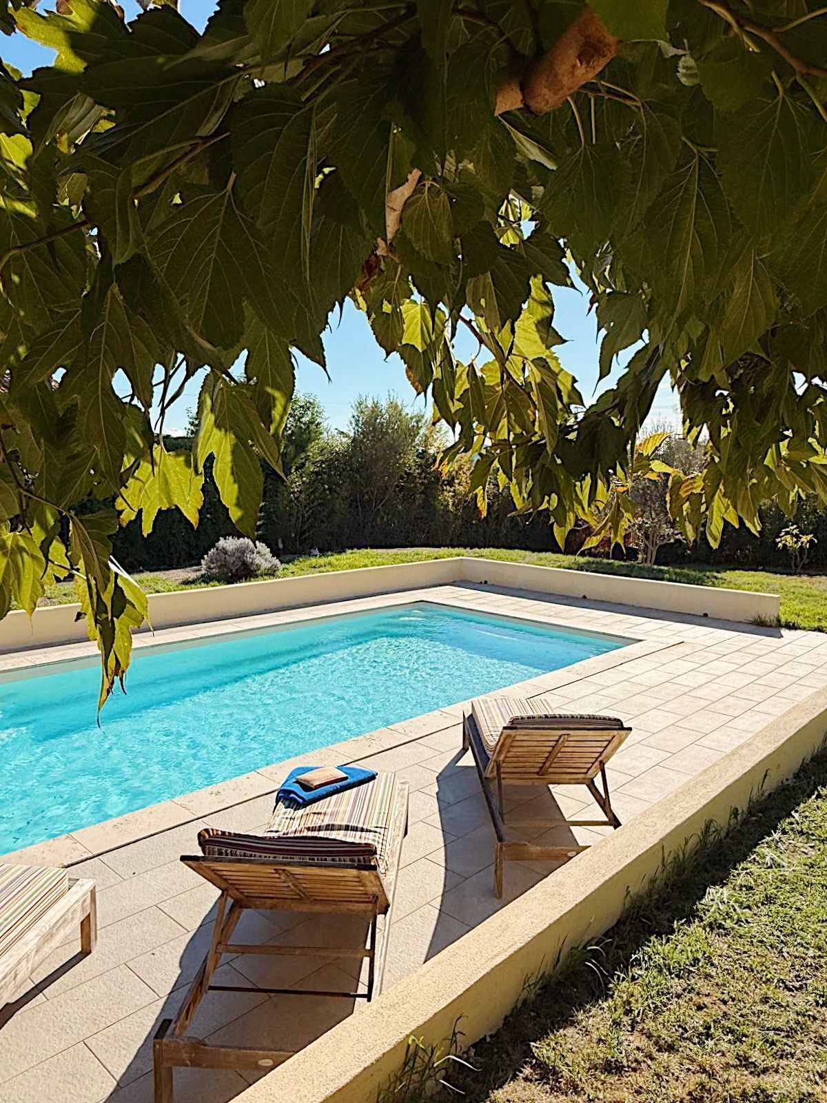Une piscine rectangulaire à l’eau d’un bleu limpide, entourée d’une terrasse carrelée. On y trouve deux chaises longues en bois et une petite table sur laquelle est posée une serviette bleue.