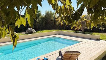 Une piscine rectangulaire à l’eau d’un bleu limpide, entourée d’une terrasse carrelée. On y trouve deux chaises longues en bois et une petite table sur laquelle est posée une serviette bleue.