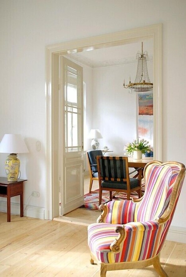 Una habitación con piso de madera, un sillón a rayas, una mesa de comedor con sillas, una araña y una puerta que da a otra habitación.