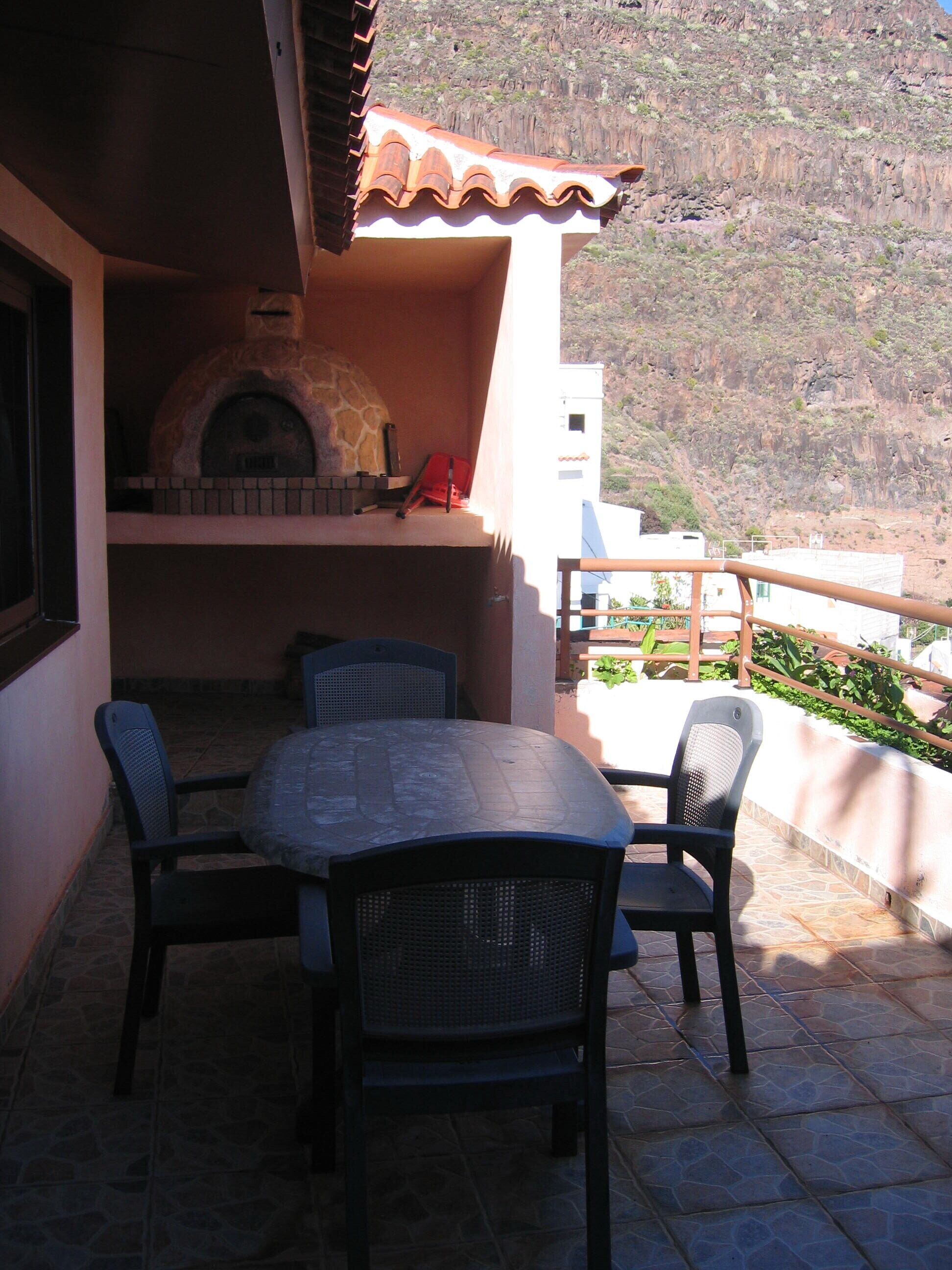 An outdoor patio with a stone table and chairs, a stone oven, and a view of a mountainous landscape.