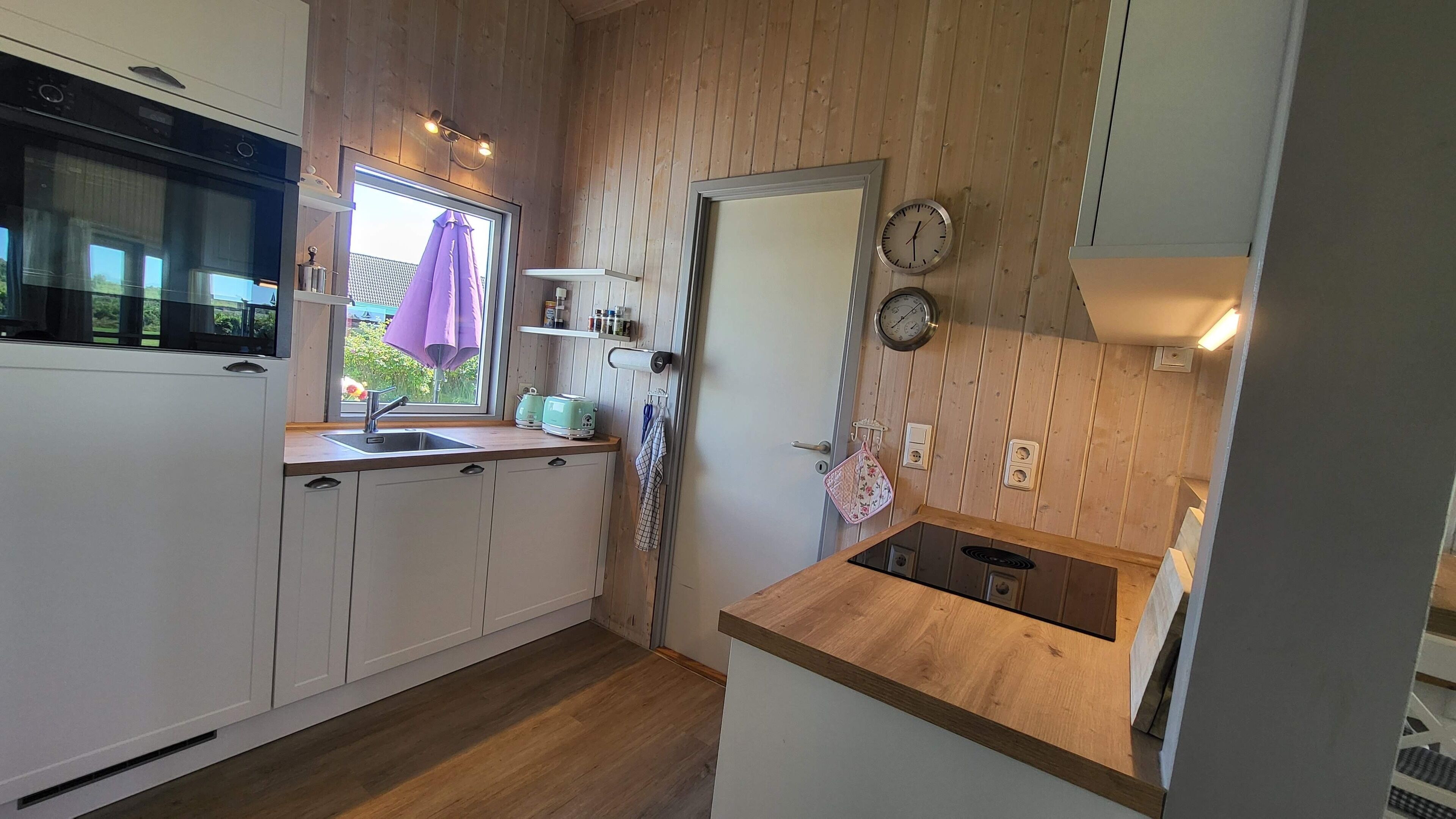 Een compacte keuken met houten wanden, een inbouwoven, een raam met een paarse paraplu en een klein spoelgedeelte.