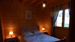 Et soveværelse i en træhytte med en seng, to sengeborde med lamper, en hængende lysekrone og et vindue med gardiner.