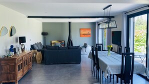 A modern living room with a sofa, dining table, and chairs. There is a fireplace, artwork on the wall, and a view of greenery outside. - Villa 220m2, land 3000m2, 5 "village on foot, 4km Saint-Rémy-de-Provence (Eyragues)