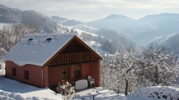 A small wooden cabin with a sloped roof, surrounded by snow-covered trees and mountains in the background.