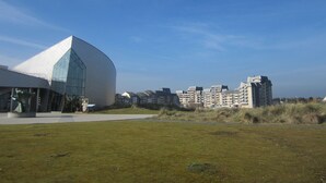 Un bâtiment moderne à l’architecture géométrique originale, entouré d’une zone verdoyante et bordé, au loin, par des immeubles d’habitation.