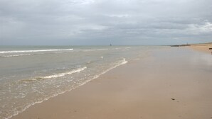 Une plage avec un phare visible au loin, des vagues calmes et un ciel nuageux.