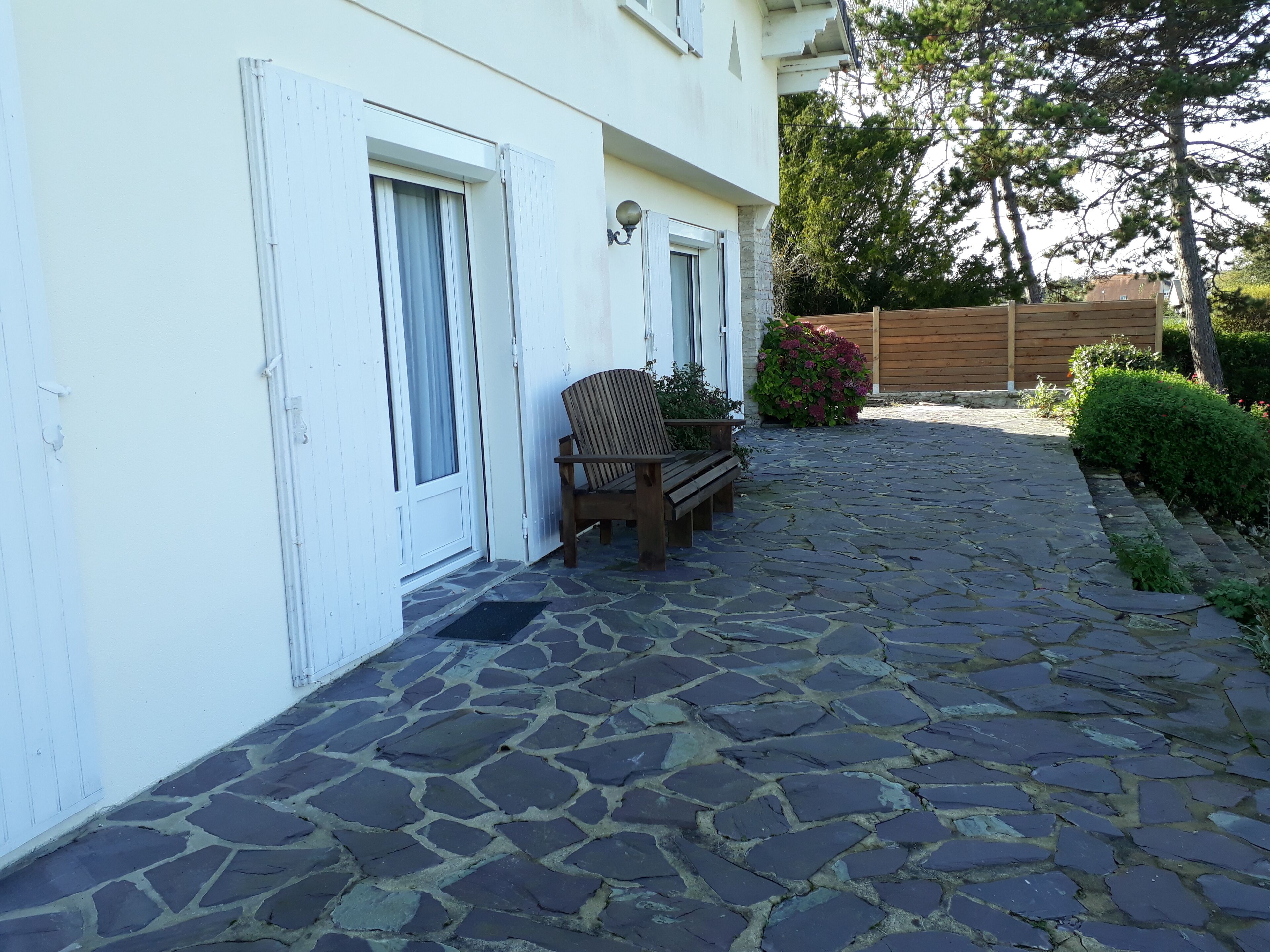 Eine terrasse mit Steinpflaster, einer hölzernen Bank und einer Tür zu einem Gebäude mit weißen Fensterläden.