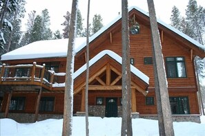 Cozy cabin with a steeply pitched roof, wooden beams, and a stone foundation, set amidst tall trees.