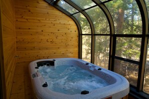 A hot tub in a wooden cabin with a curved glass window offering a forest view.