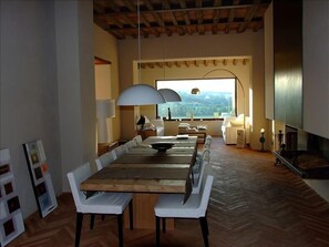 Enjoy meals in this spacious dining area with a long table and comfortable chairs, surrounded by a high ceiling and large window offering a view of the outdoors.
