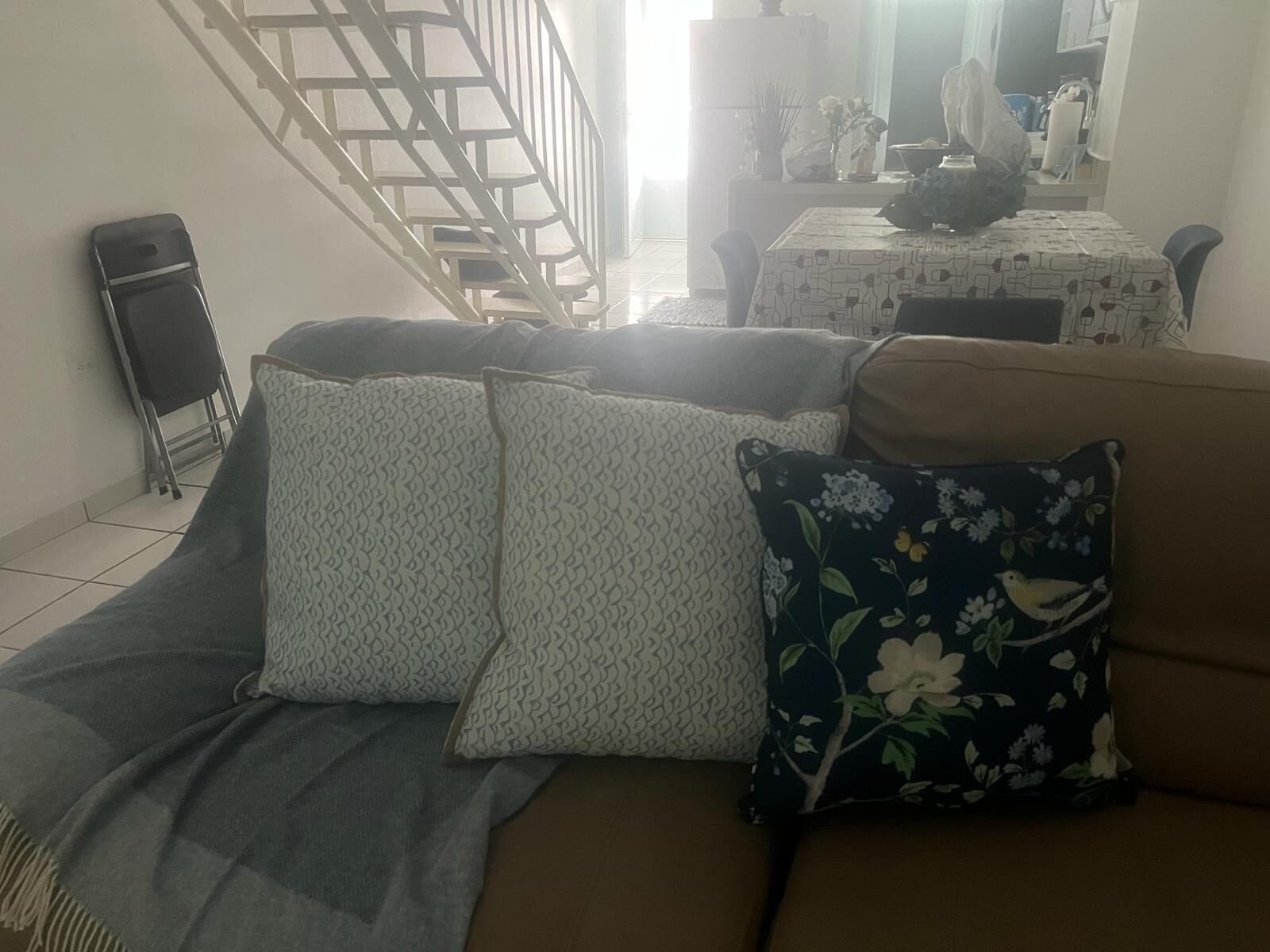 A living room with a sofa, patterned cushions, a small table with a vase, and a staircase in the background.