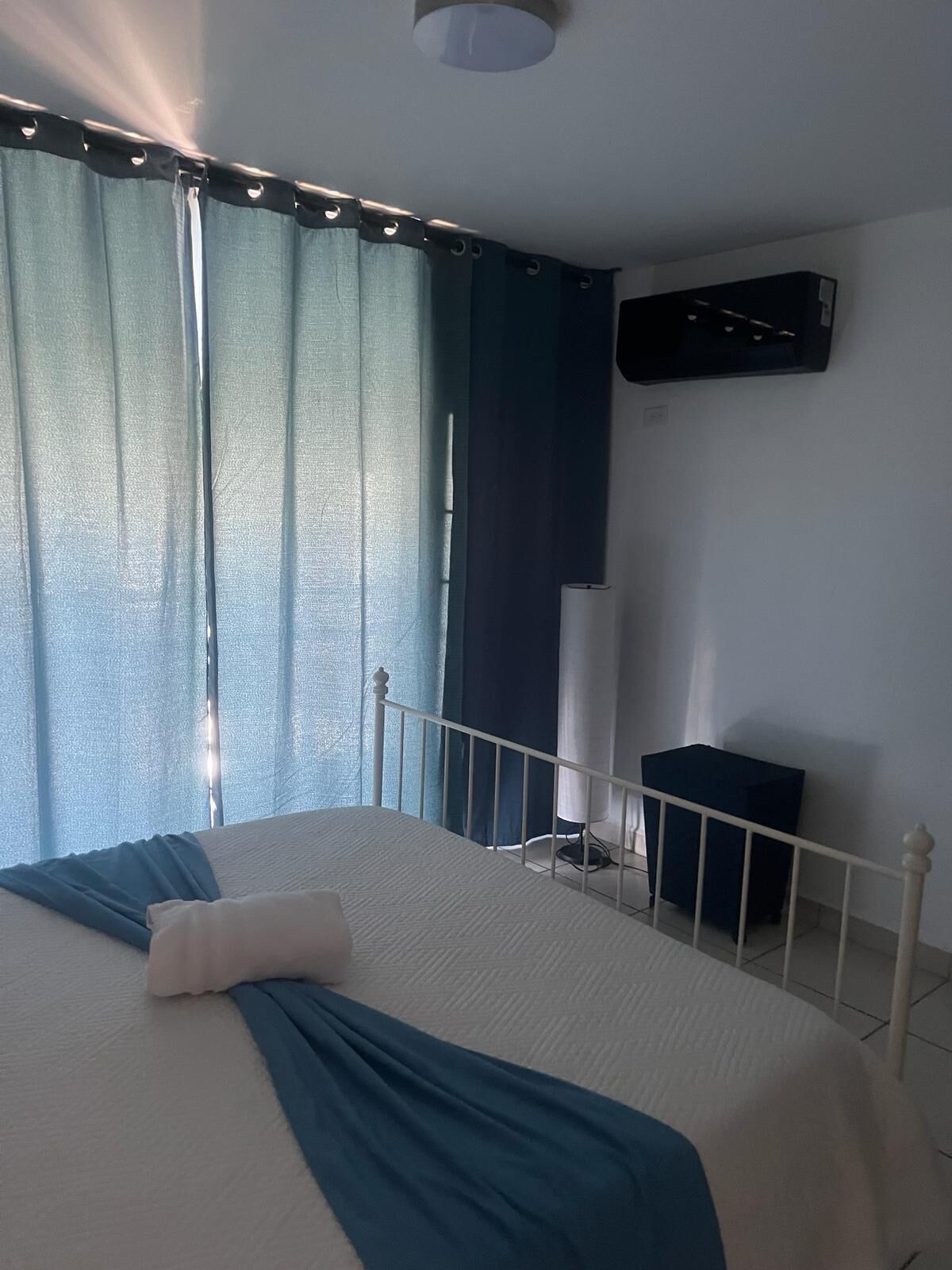 A bedroom with a bed, curtains, a bedside table, and a wall-mounted air conditioner.